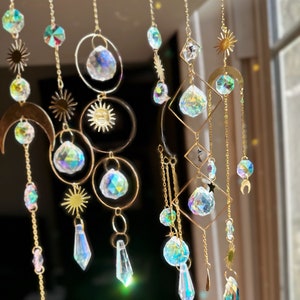 May include: A gold metal suncatcher with multiple crystal prisms hanging from it. The suncatcher features a crescent moon, a sun, and a star design. The crystals are iridescent and reflect light in a rainbow of colors.