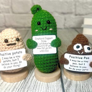 May include: Three crocheted figures: a potato, a pickle, and a poo. Each figure holds a sign with a positive message. The potato is tan, the pickle is green, and the poo is brown. They sit on small wooden bases.