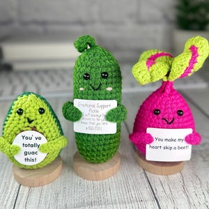 May include: Three crocheted food-themed plushies: an avocado, a pickle, and a beet. The avocado is green and says "You've totally guac this!". The pickle says "Emotional Support Pickle" and "a BIG DILL!". The beet says "You make my heart skip a beet!"