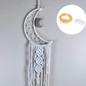 May include: White crescent moon dream catcher with a silver crescent moon charm hanging from the bottom. The dream catcher has a white macrame design and long white fringe. A string of white fairy lights is shown in the background.