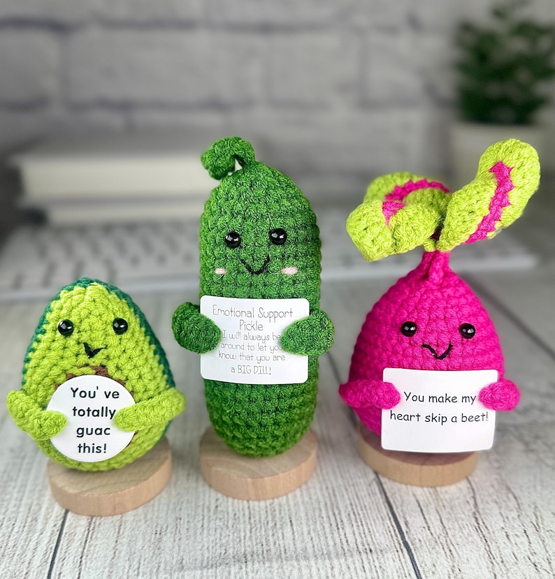 May include: Three crocheted food-themed plushies: an avocado, a pickle, and a beet. The avocado is green and says "You've totally guac this!". The pickle says "Emotional Support Pickle" and "a BIG DILL!". The beet says "You make my heart skip a beet!"
