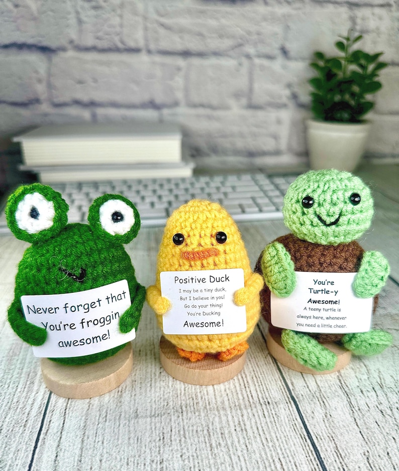 May include: Three crocheted amigurumi figures: a green frog, a yellow duck, and a green turtle. Each holds a sign with an inspirational message. The frog says, "Never forget that You're froggin awesome!" The duck says, "Positive Duck...You're Ducking Awesome!" The turtle says, "You're Turtle-y Awesome!"