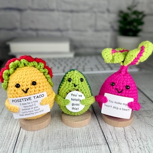 May include: Three crocheted food-themed figures: a yellow taco with red and green trim, a green avocado, and a pink beet. Each figure holds a small sign with a pun. They are displayed on wooden bases, with a blurred background.
