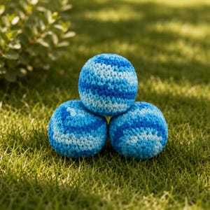May include: Three spherical, hand-crocheted balls arranged in a pyramid formation. Each ball features a variegated pattern of yarn in shades of dark blue, medium blue, and light blue, creating a textured, soft surface with a consistent stitch.