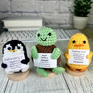 May include: Three crocheted figurines: a penguin, a turtle, and a duck. The penguin is black and white, the turtle is green and brown, and the duck is yellow and orange. Each holds a sign with an inspirational message. They are mounted on small wooden bases.