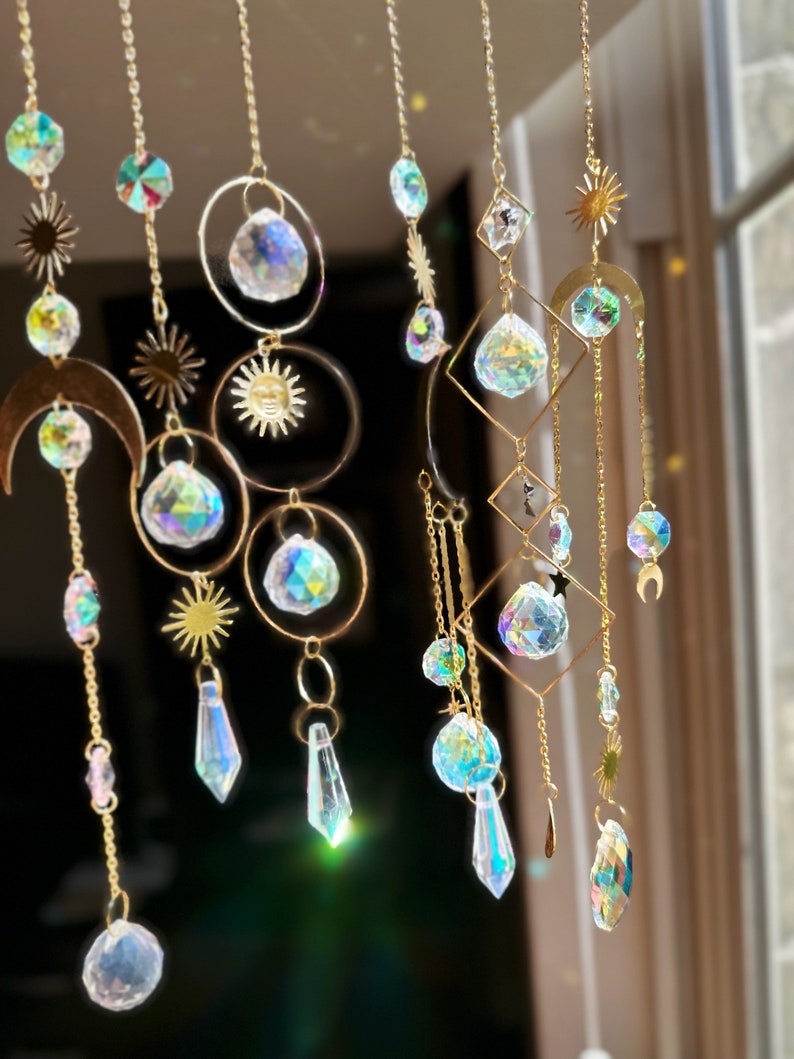 May include: A gold metal suncatcher with multiple crystal prisms hanging from it. The suncatcher features a crescent moon, a sun, and a star design. The crystals are iridescent and reflect light in a rainbow of colors.