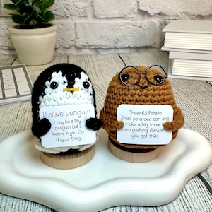 May include: Two crocheted plushies, a penguin and a potato, holding signs with positive messages. The penguin's sign reads "Positive penguin I may be a tiny penguin, but I believe in you Go do your thing!" The potato's sign reads "Cheerful Potato Small potatoes can still make a big impact Keep pushing forward. you got this!"