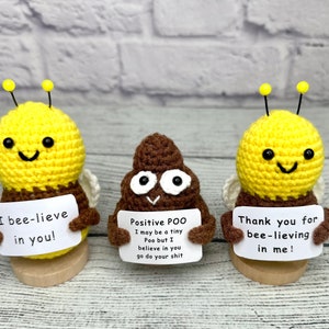 May include: Three handmade crochet figures: two yellow and brown bees and a brown poop emoji. Each holds a sign with a positive message. The bees have yellow antennae and white wings. The poop emoji has a smiling face.