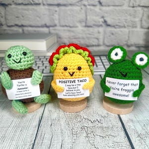 May include: Three crocheted amigurumi figures: a turtle, a taco, and a frog. Each holds a sign with an encouraging message. The turtle is brown and green, the taco is yellow with red and green accents, and the frog is green.