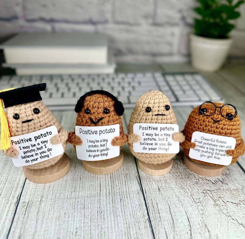 May include: Four crocheted potato figures, each holding a sign with an inspirational message. One wears a graduation cap, another has headphones, a third has glasses. The potatoes are on wooden bases.
