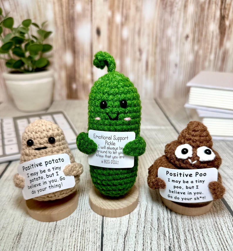 May include: Three crocheted figures: a tan potato, a green pickle, and a brown poo. Each holds a sign with a positive message. The potato says "Positive potato," the pickle says "Emotional Support Pickle," and the poo says "Positive Poo."