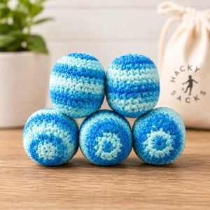 May include: Five round, crocheted hacky sacks featuring a variegated pattern of light blue, medium blue, and dark blue yarn. The spheres are arranged in a pyramid formation, with a beige drawstring bag labeled HACKY SACKS in the background.