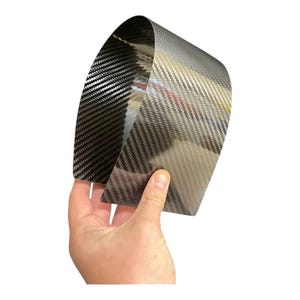 Puede incluir: Un trozo de material de fibra de carbono se sostiene sobre un fondo blanco. El material es de color gris oscuro con un patrón tejido. El material está curvado, mostrando su flexibilidad. La imagen está bien iluminada, resaltando la textura y los detalles de la fibra de carbono.