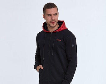 Può includere: Felpa blu navy con zip e fodera del cappuccio rossa. La felpa presenta un piccolo logo sulla manica sinistra e testo rosso sul petto. Il modello indossa jeans grigio scuro.