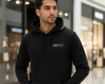 May include: Black hoodie with a front pocket and a drawstring hood. The hoodie has white text on the chest and a striped design on the hood's edge. The person is wearing black jeans. The background is a blurred indoor shopping area.