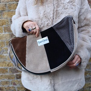May include: A beige, brown, and black corduroy crescent-shaped bag with a black leather strap. The bag has a label that says "Crafty Threads".