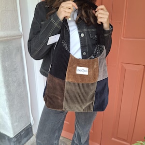 Op de afbeelding: Een patchwork corduroy tote bag in bruin, zwart en marineblauw. De tas heeft een label met de tekst "CAMDEN THREADS". De tas wordt vastgehouden door een persoon die een zwarte spijkerjas en een grijze jeans draagt. De tas is ongeveer 35 cm breed.
