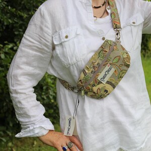 May include: A beige fanny pack featuring a multi-colored floral and leaf pattern in shades of green, orange, and brown. The bag has a zipper closure, an adjustable strap, and a white rectangular label with the text Queen Threads attached.