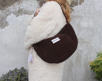 May include: A brown corduroy shoulder bag with a curved shape. The bag has a white label with text on it. The bag is worn over the shoulder. The background is a textured wall and some greenery.