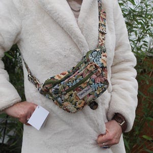 Puede incluir: Un bolso de cintura de tapiz floral con cremallera y correa verdes. La bolsa presenta un colorido estampado de flores y hojas. La correa es ajustable y la bolsa se lleva cruzada. La etiqueta dice "Fabric Threads".