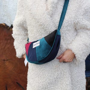 以下が含まれることがあります： ティールストラップが付いた三日月型のショルダーバッグ。バッグは、バーガンディ、ブラック、ティール、ネイビーブルーのパネルが特徴です。テキスト付きの白いタグが添付されています。バッグは肩からかけています。