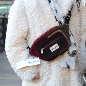 Puede incluir: Riñonera de pana multicolor con cierre de cremallera y una pequeña etiqueta que dice "Timber Threads". La bolsa es una mezcla de borgoña, marrón y verde oliva. La bolsa se lleva sobre un abrigo blanco y esponjoso.