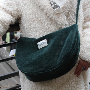 Könnte beinhalten: Dunkelgrüne Cord-Umhängetasche mit geschwungener Form. Die Tasche hat einen langen Riemen und ein kleines rechteckiges Etikett mit dem Text "TinkerThreads". Die Tasche wird über einem weißen, flauschigen Mantel getragen.