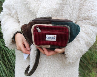 May include: A corduroy fanny pack in shades of brown, burgundy, and green. The pack has a small zippered pocket on the front with a label that reads "Camden Threads". The bag is held by a person wearing a white, fluffy coat.