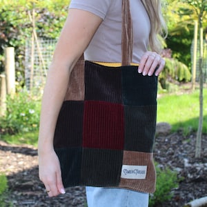 Puede incluir: Una bolsa de mano de patchwork hecha de pana en tonos marrón, verde y burdeos. La bolsa tiene un forro amarillo y una etiqueta de cuero marrón con el texto "Camden Threads".