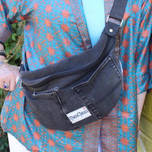 Op de afbeelding: Een zwarte crossbodytas van denim met een gebogen ritssluiting, een buitenzak en een wit rechthoekig label met de tekst Camden Threads. De tas heeft een brede zwarte stoffen riem en is gemaakt van gerecyclede donkere denimstof.
