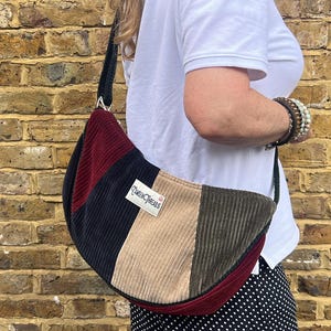 May include: A corduroy shoulder bag with a patchwork design in burgundy, navy, tan, and olive green. The bag has a black strap and a small rectangular tag with text. The bag is worn over the shoulder.