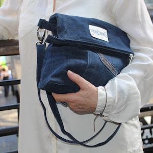 May include: A dark blue denim fold-over crossbody bag featuring a silver metal zip closure, a matching denim shoulder strap with a silver swivel clasp, and a white rectangular fabric label with the text "Urban Threads" stitched on the front.