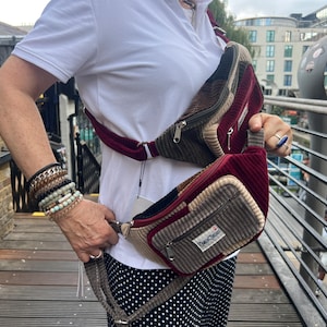 Puede incluir: Riñonera de pana en burdeos, marrón y beige. La bolsa tiene múltiples bolsillos y un compartimento principal con cremallera. El bolsillo delantero tiene una etiqueta que dice "Outer Classic". La correa es ajustable.