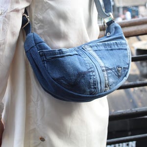 May include: A crescent-shaped crossbody bag constructed from blue denim fabric. The bag features visible stitching, a front pocket with a triangular logo patch, and a wide denim shoulder strap attached with silver-toned metal hardware rings.