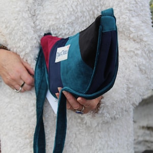 May include: A multi-coloured shoulder bag with a teal, navy, burgundy, and black design. The bag has a long teal strap and a label that reads "Dust Clouds". The bag is held by a person wearing a white fuzzy coat.