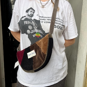 May include: A corduroy shoulder bag with a patchwork design in burgundy, black, brown, and olive green. The bag has a curved shape and a long strap. The bag has a tag that says "Camden Threads". The person is wearing a white t-shirt with a graphic print.