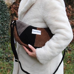 May include: A corduroy shoulder bag with a patchwork design in shades of brown, black, and gray. The bag has a black strap and a label that reads "TANGEN THREADS". The bag is held by a person wearing a white coat.
