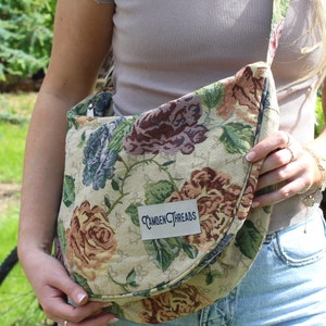Könnte beinhalten: Schultertasche mit Blumenmuster mit cremefarbenem Hintergrund und großen Rosen in Braun-, Blau- und Grüntönen. Die Tasche hat eine gebogene Klappe und einen Riemen. Auf dem Etikett steht "Camden Threads".