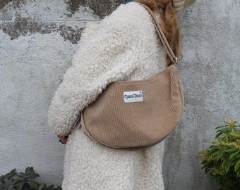 May include: A tan corduroy shoulder bag with a curved shape and a long strap. The bag has a rectangular label with the text "Three Cheers". The bag is worn over a white, fluffy coat.