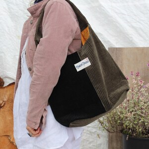 Puede incluir: Un bolso tote de pana en verde oliva, negro y naranja. El bolso se lleva al hombro. Una etiqueta blanca con el texto "Queen Classics" está cosida en la parte delantera. El bolso forma parte de una colección de bolsos hechos a mano.