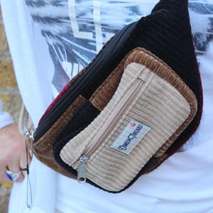 May include: A multi-colored corduroy fanny pack with a zipper closure. The bag features a combination of black, brown, tan, and burgundy panels. A small rectangular label with the text "Camden Threads" is visible on the front.