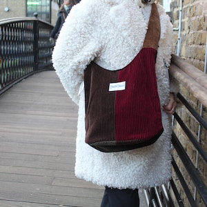 May include: A corduroy tote bag in shades of brown and burgundy, with a rectangular label that reads "Dawn Chorus". The bag has a rounded bottom and a long strap. The person is wearing a white, fluffy coat.
