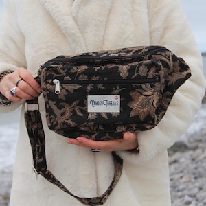 May include: A black fanny pack with a floral pattern in shades of brown and gold. The bag has a zippered main compartment and a front pocket. The strap is adjustable. The label reads "Camden Threads".