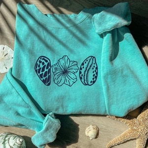 May include: A light teal sweatshirt featuring a dark blue seashell and flower design. The sweatshirt is folded on a light wood surface, with a starfish, seashells, and a sand dollar nearby. The design includes a seashell, a flower, and another seashell.