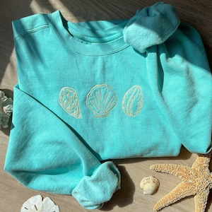 May include: A light blue sweatshirt with three embroidered seashells in a row. The seashells are in a light tan color. The sweatshirt is on a wooden surface with a starfish and seashells.