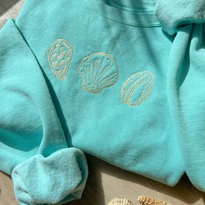 May include: A light blue sweatshirt with embroidered seashell designs in cream. The crew neck sweatshirt has long sleeves with ribbed cuffs. A seashell and starfish are on the wooden surface.