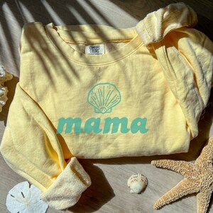 May include: A light yellow sweatshirt with a seashell graphic and the word "mama" in a teal color. The sweatshirt is made of a soft material and has a crew neck. The sleeves are rolled up.