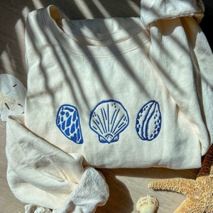 May include: Cream-colored sweatshirt featuring three blue embroidered seashell designs. The sweatshirt has a crew neck and long sleeves. The seashells are a scallop, a clam, and a conch. The sweatshirt is made of a soft, comfortable material.