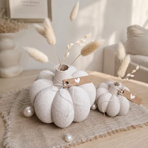 May include: Two off-white, textured, pumpkin-shaped ceramic vases with ribbed surfaces. Each vase features a small brown paper tag with a white heart icon, tied with thin white string, and contains dried beige grass stems and white pearls.