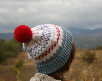 May include: A knitted winter hat with a red pom-pom. The hat features a white base with a blue and red geometric pattern and a blue ribbed brim. The hat is worn outdoors with a blurred background of a field and mountains.